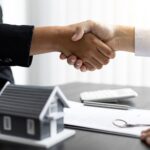 Selling Luxury Real Estate to Wealthy Clients: An Insider's Guide. Two individuals shaking hands over a table with documents, keys, and a miniature house figure on it.